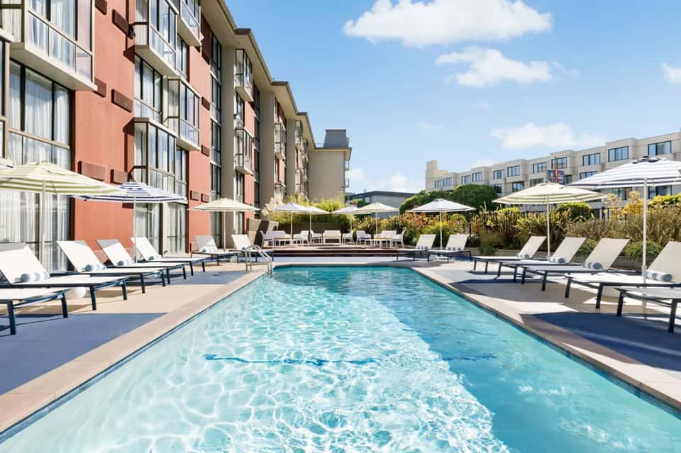 Rectangular outdoor pool with clear turquoise water, white lounge chairs, and umbrella seating surrounded by modern buildings