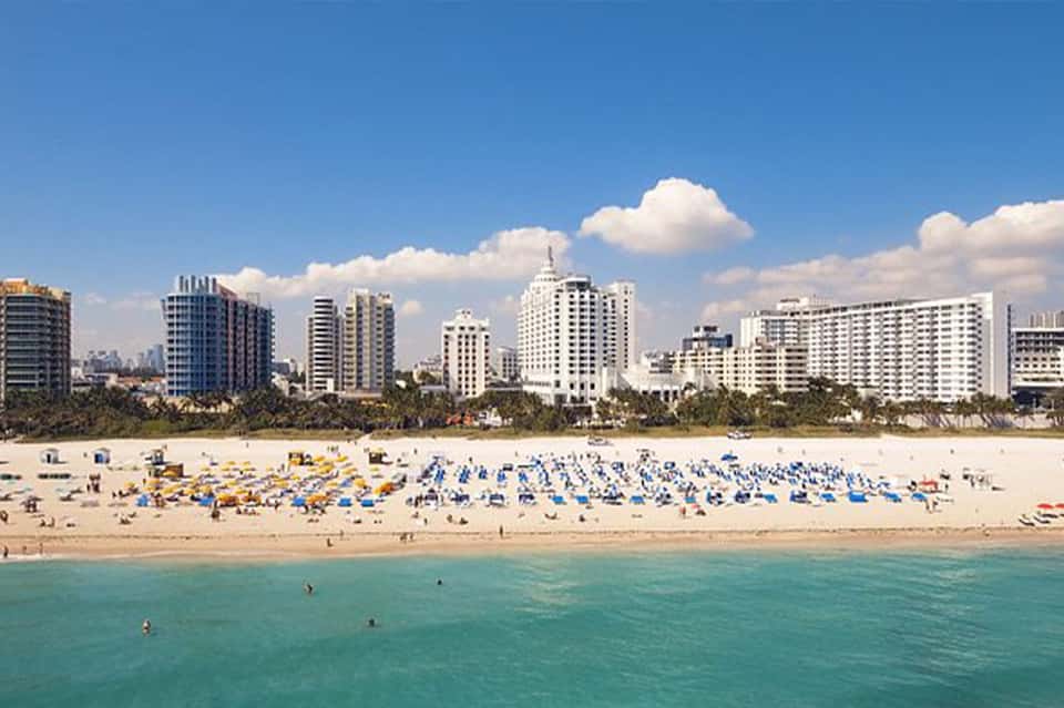 Miami Beach shoreline with colorful beach umbrellas, sunbathers, and high-rise hotels along sandy coast