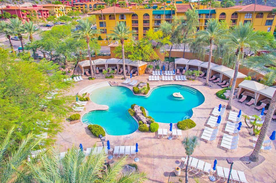 Aerial view of resort with turquoise pool, palm trees, colorful buildings, and lounge chairs surrounding the pool deck