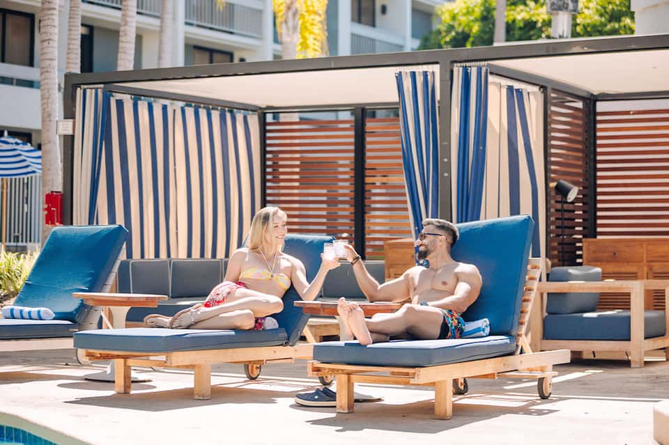 Couple relaxing on wooden lounge chairs with blue cushions under striped cabana by resort pool