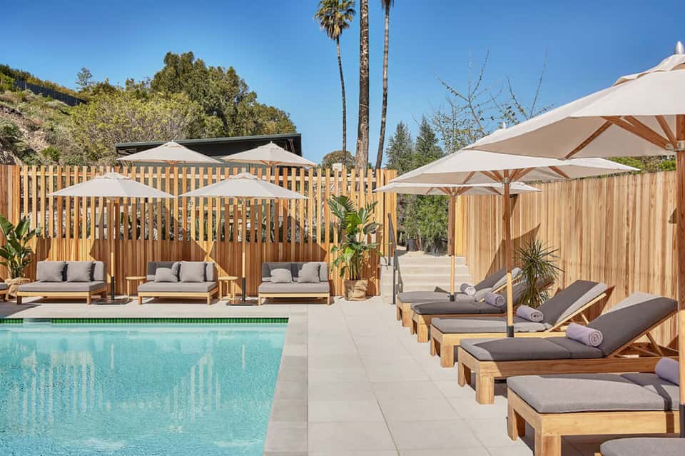 Resort pool area with wooden loungers, white umbrellas, palm trees, and hillside views