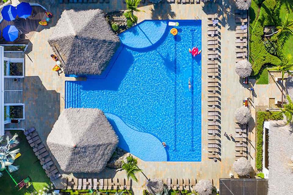 Aerial view of curved resort pool with thatched umbrellas, lounge chairs, and surrounding grounds