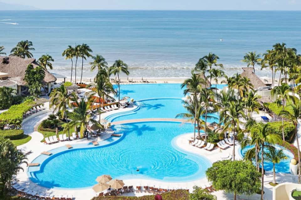 Large resort pool complex with curved sections, palm trees, beach, and ocean in background