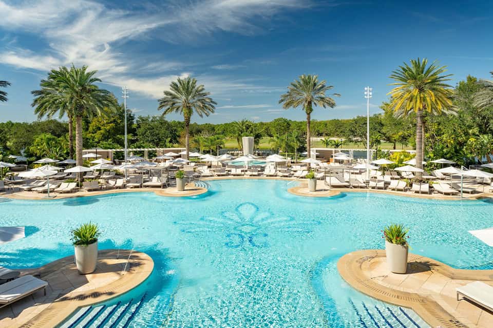 Resort pool area with crystal-clear turquoise water, loungers, palm trees, and white umbrellas on sunny day