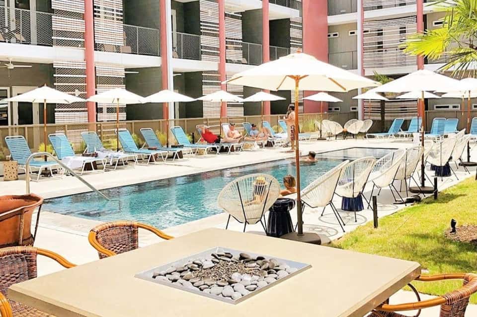 Pool area with fire pit, white umbrellas, lounge chairs, and colorful modern building with balconies
