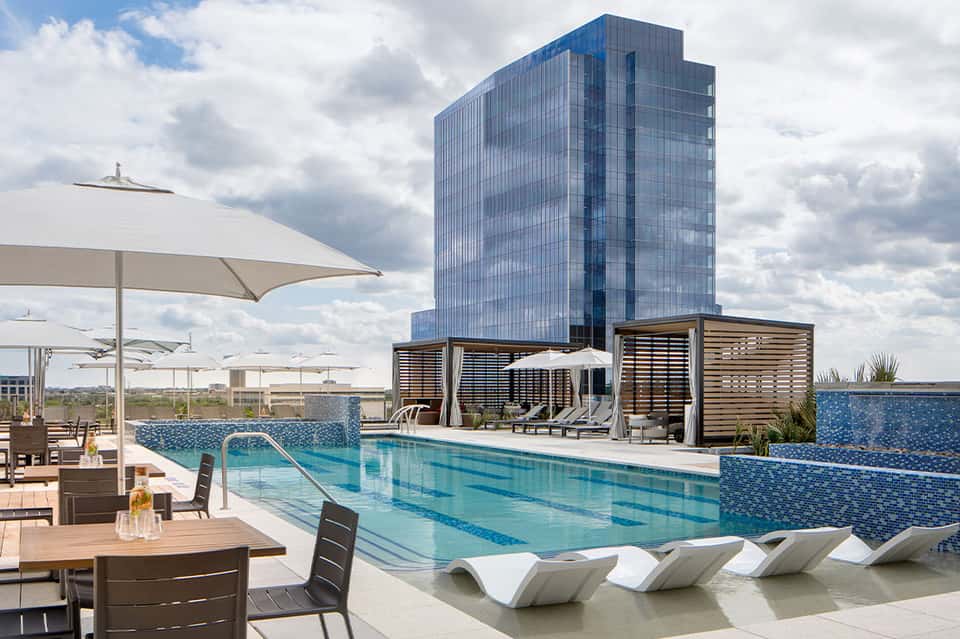 Modern hotel rooftop pool with lap lanes, loungers, glass tower building, and white umbrellas