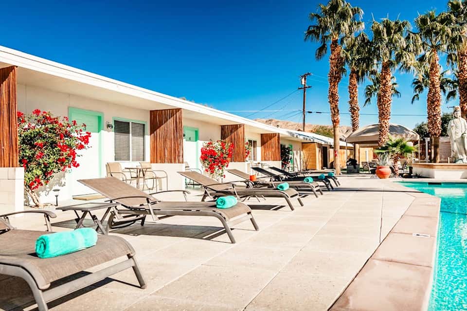 Resort poolside with turquoise water, lounge chairs, palm trees, and white bungalows with red flowers
