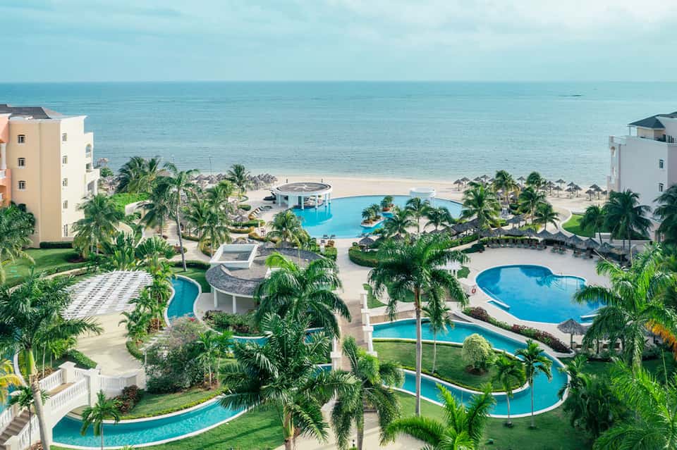 Beachfront resort aerial view showing multiple pools, palm trees, sandy beach, and ocean with residential buildings