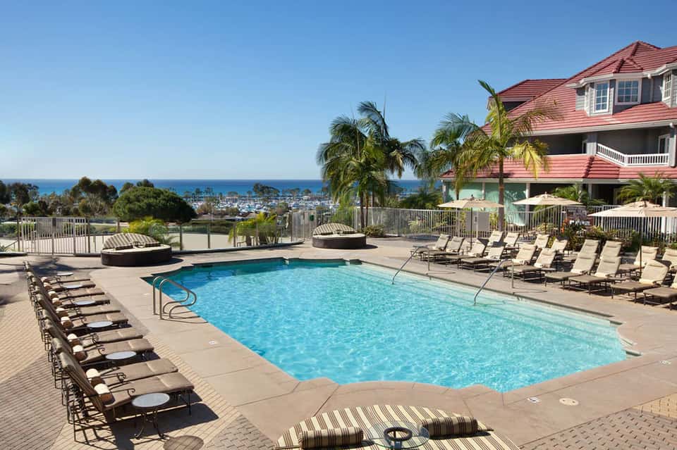 Resort pool overlooking coastal town with palm trees, lounge chairs, and distant ocean view