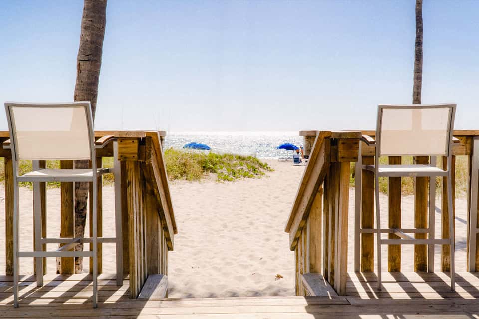Wooden beach chairs with canvas backs facing sandy beach and ocean with palm trees
