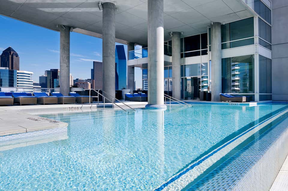 Indoor lap pool with lane markings, concrete columns, city skyline views, and blue lounge seating area