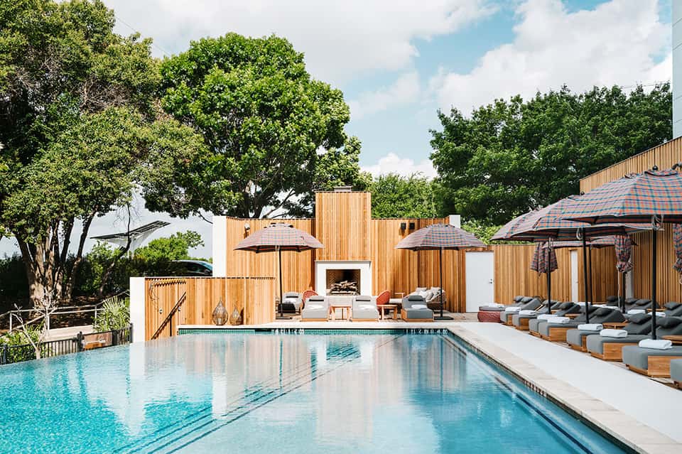 Modern resort pool with wooden architecture, lounge chairs, umbrellas, and mature trees