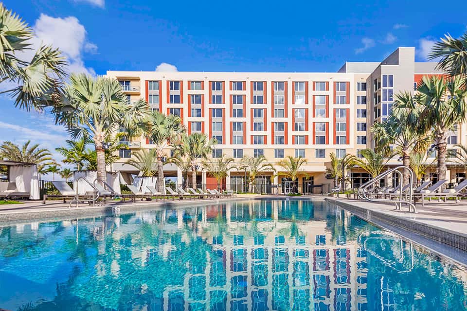 Modern resort with orange and white striped facade, palm trees, and crystal-clear pool under blue sky