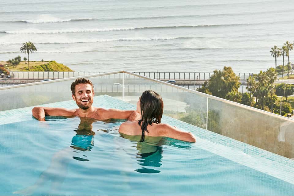 Couple relaxing in outdoor infinity pool overlooking beach and ocean with glass railings and palm trees