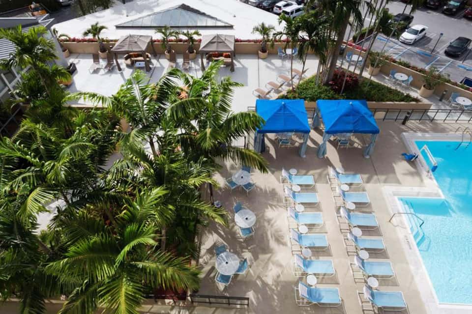Aerial view of resort pool area with blue umbrellas, lounge chairs, palm trees, and surrounding amenities
