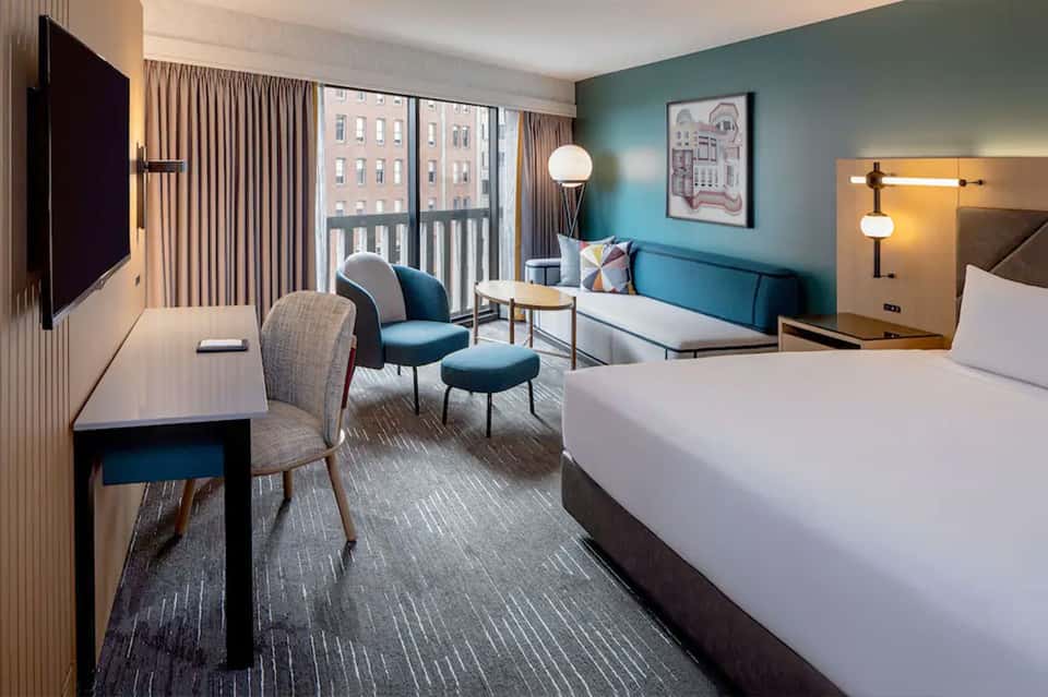 Modern hotel suite with bed, teal accent wall, lounge chair, and city views