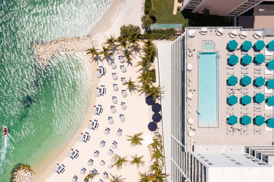 Beach resort aerial view with infinity pool, lounge seating, turquoise umbrellas, and ocean waterfront
