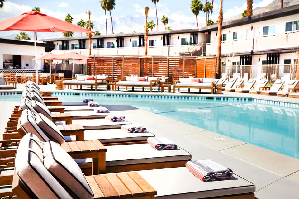 Resort pool deck with wooden lounge chairs, red umbrellas, and palm-lined grounds