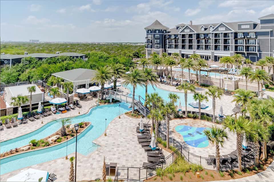 Aerial view of curved resort pool with palm trees, multi-story hotel, and waterfront location