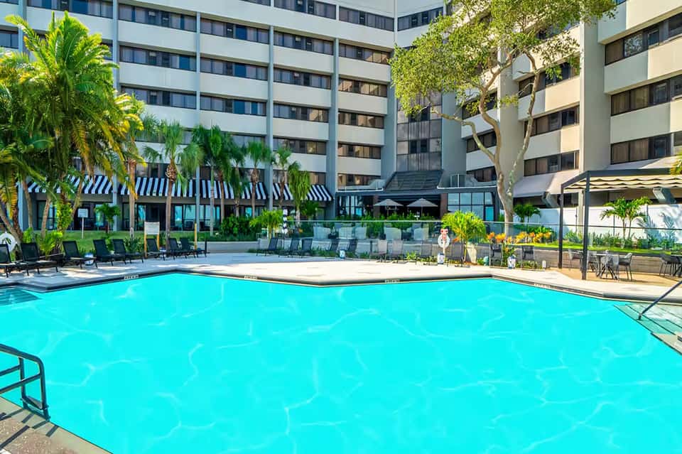 Large resort pool surrounded by palm trees, striped umbrellas, and multi-story hotel complex