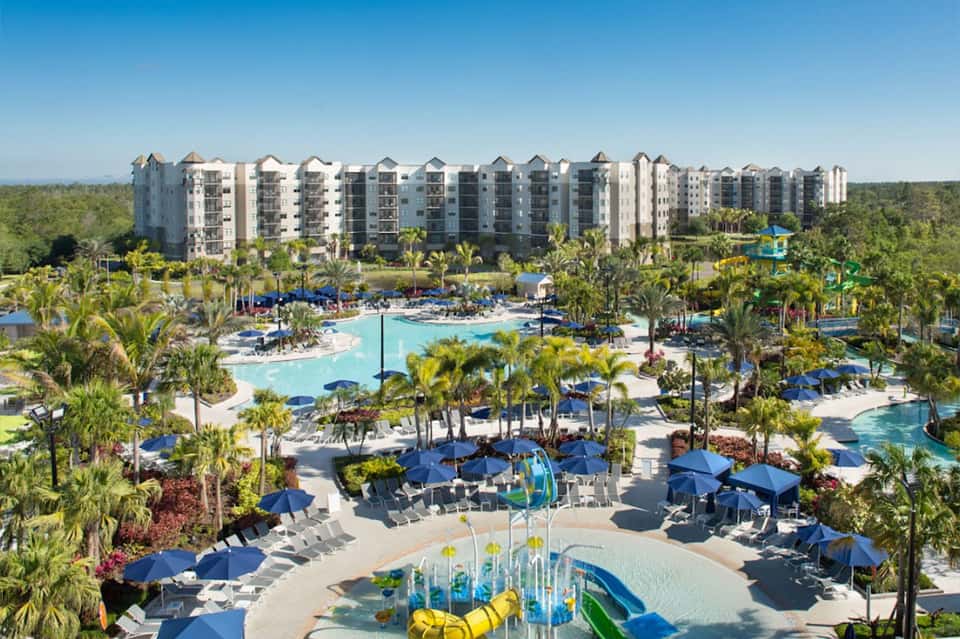 Aerial view of large resort complex with multiple swimming pools, water features, palm trees, and residential towers