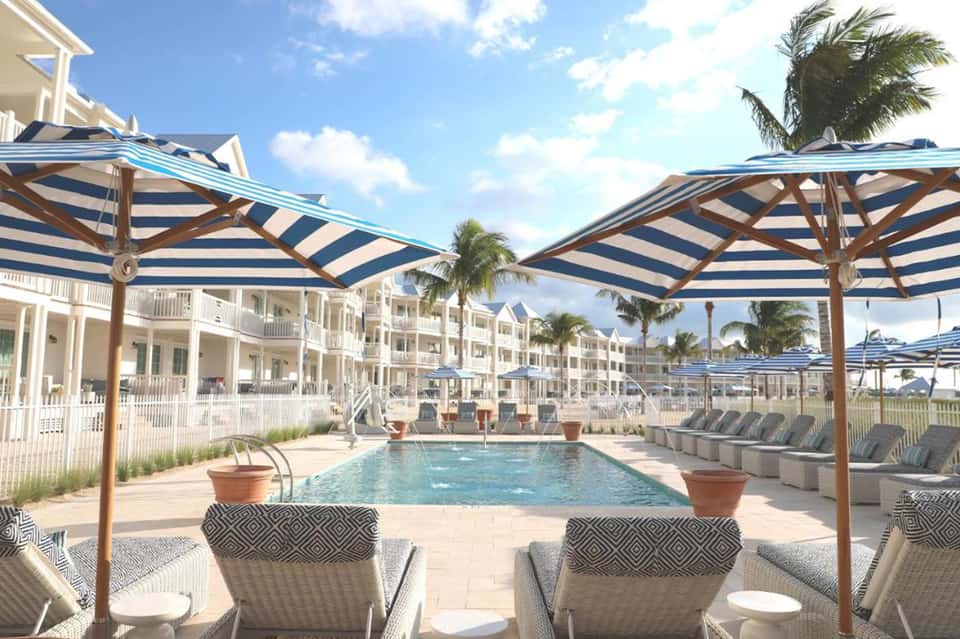 Resort pool area with striped umbrellas, loungers, white residential buildings, and palm trees