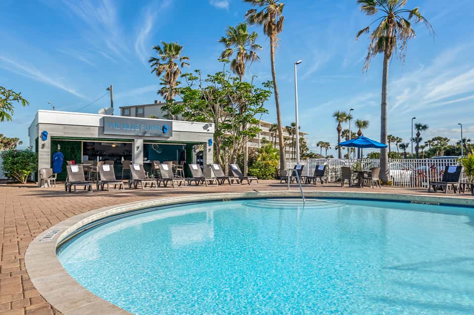 Resort pool area with blue umbrellas, lounge chairs, and beachside bar under palm trees