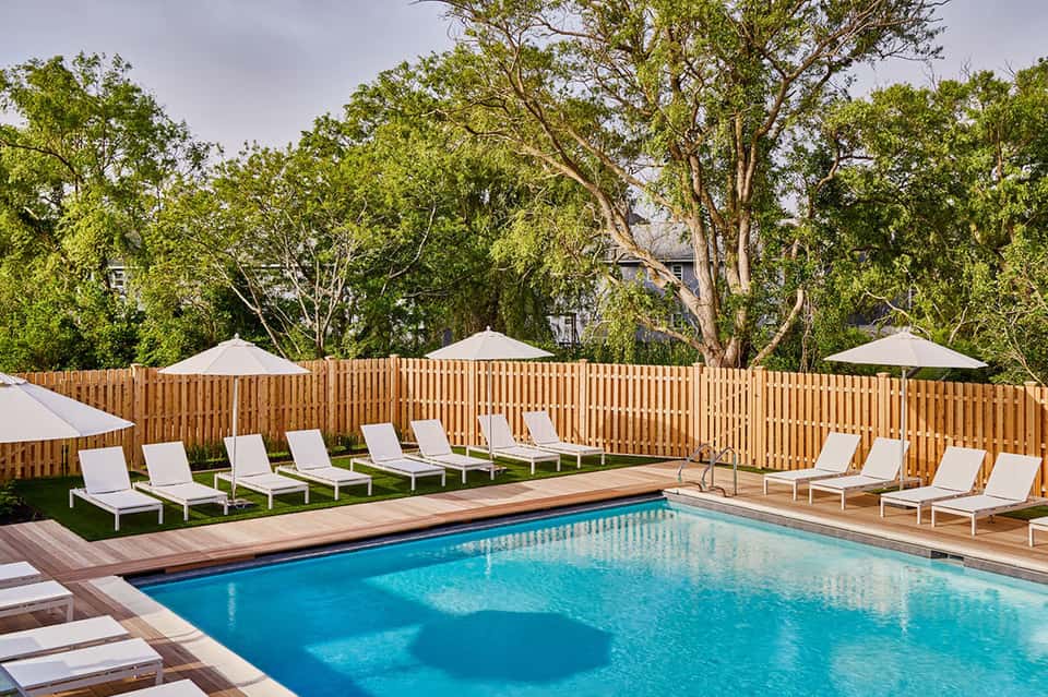 Resort pool with loungers, umbrellas, wooden fence, and surrounding trees