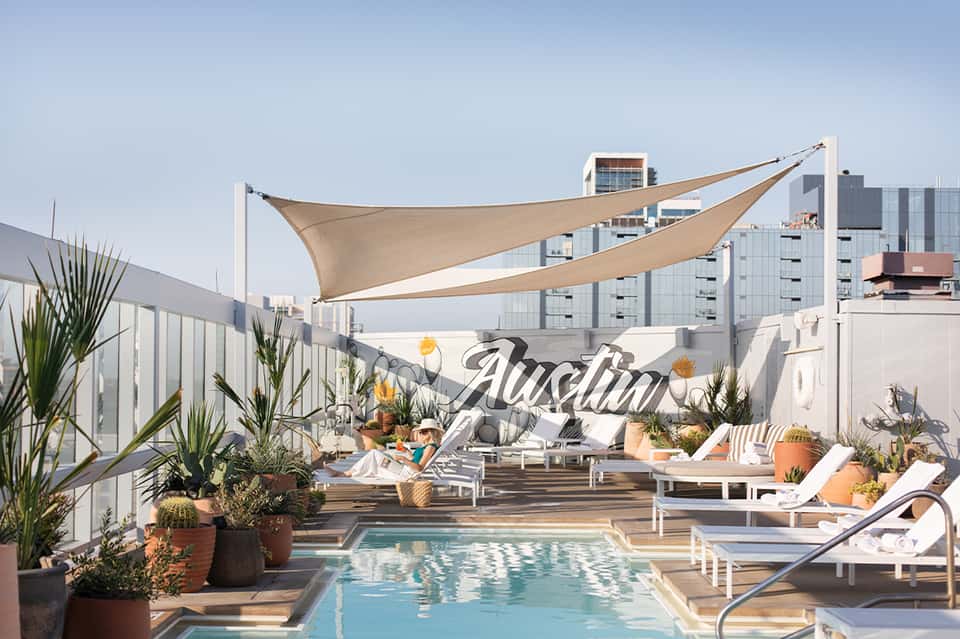 Rooftop pool deck with beige sail shade, loungers, potted plants, and urban skyline backdrop