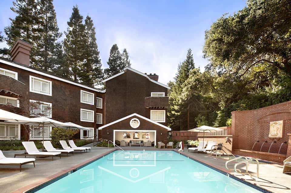 Resort exterior with brick buildings, tall evergreen trees, lounge chairs, and crystal-clear swimming pool