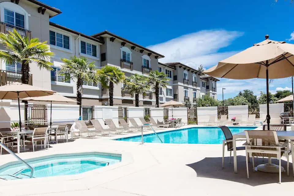 Resort pool area with loungers, shade umbrellas, spa tub, and multi-story hotel building