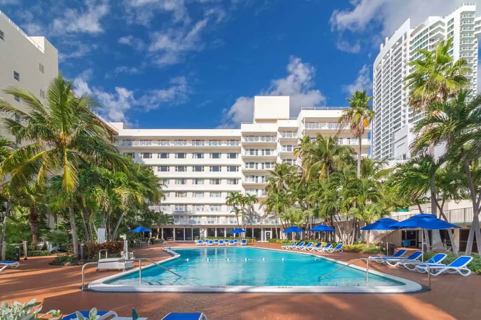 Resort courtyard pool surrounded by multi-story hotel buildings, palm trees, and blue umbrellas