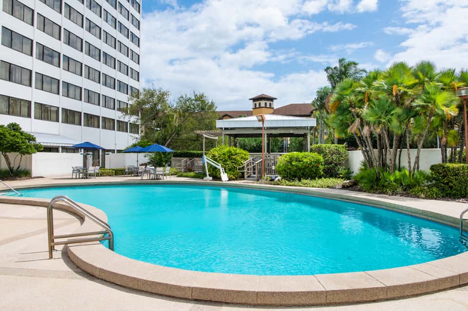 Curved turquoise pool with palm trees, gazebo, white hotel building, and lounge areas