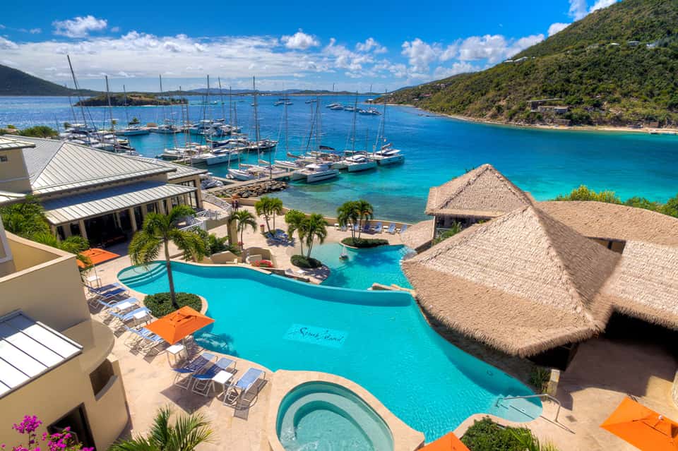 Aerial view of beachfront resort pool with thatched bungalows, boats, and turquoise bay
