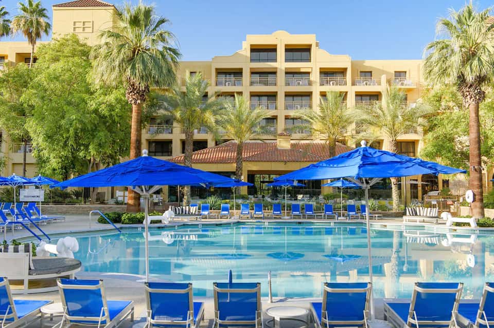 Resort pool with bright blue umbrellas, loungers, palm-lined courtyard, and multi-story yellow building