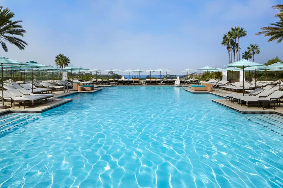 Large resort pool with crystal-clear blue water, lounge chairs, umbrellas, and palm trees on sunny day