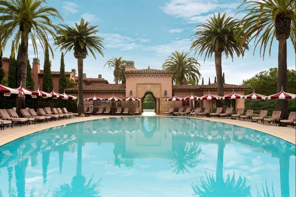 Luxurious pool surrounded by palm trees, red and white umbrellas, lounge chairs, and Moroccan architecture