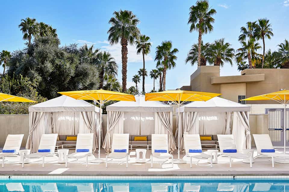 Poolside cabanas with white curtains and yellow umbrellas surrounded by palm trees