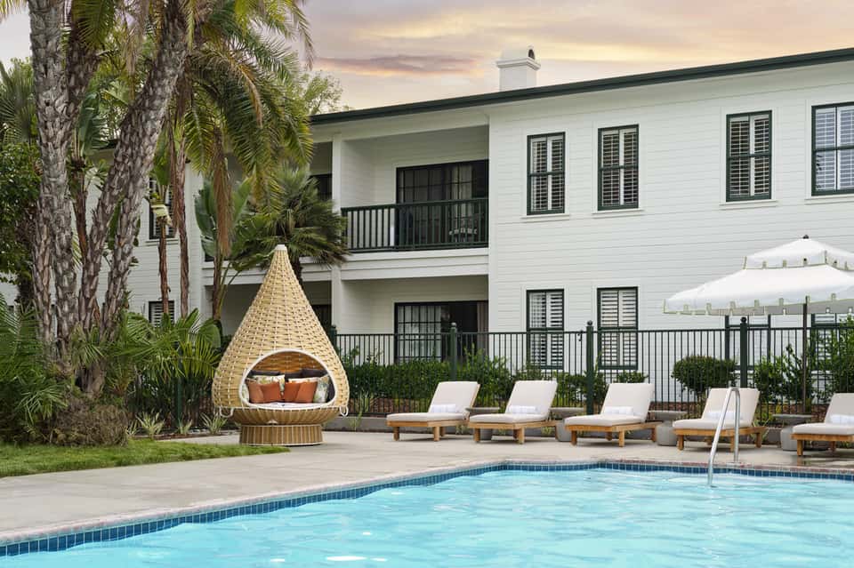 Luxury pool area with woven lounger, umbrella, palm trees, and white house backdrop
