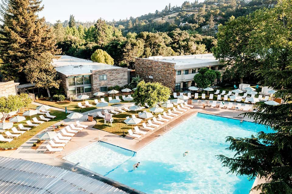 Resort pool area with lounge chairs, umbrellas, and stone building nestled in forest