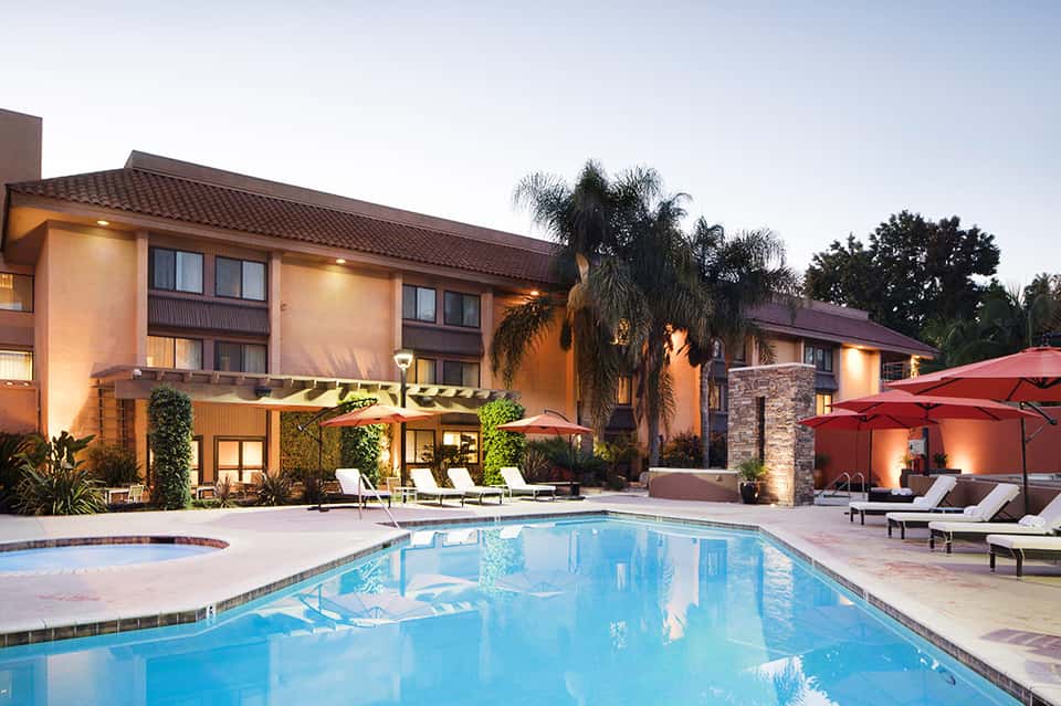 Evening resort exterior with illuminated buildings, pool, red umbrellas, and tropical landscaping