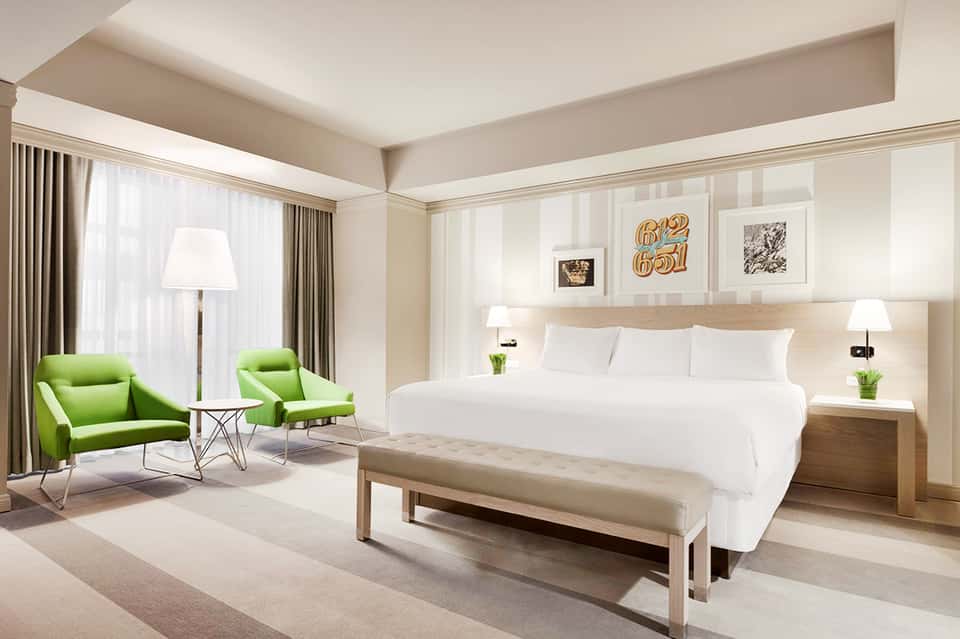 Luxury hotel suite with king bed, green lounge chairs, floor-to-ceiling windows, and contemporary light wood furniture