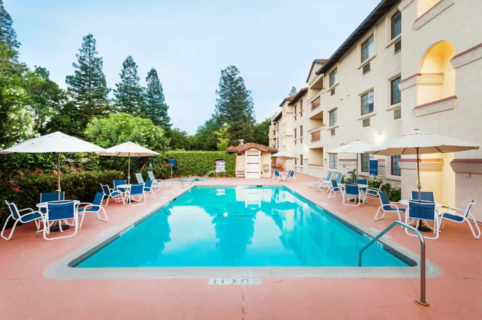 Resort pool surrounded by lounge chairs with blue cushions, white umbrellas, and residential building with arched windows
