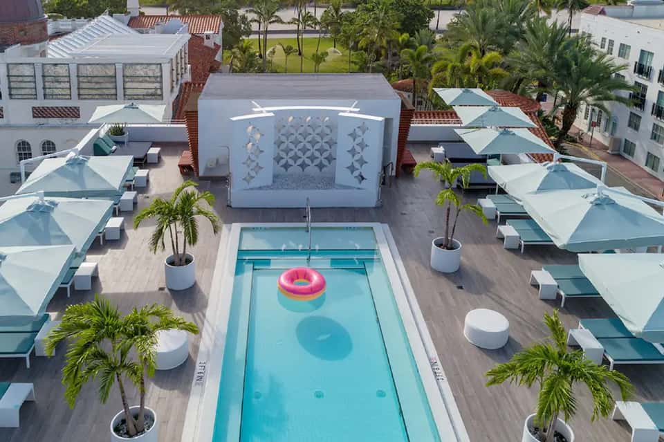 Aerial view of modern resort pool deck with blue water, white umbrellas, and surrounding buildings