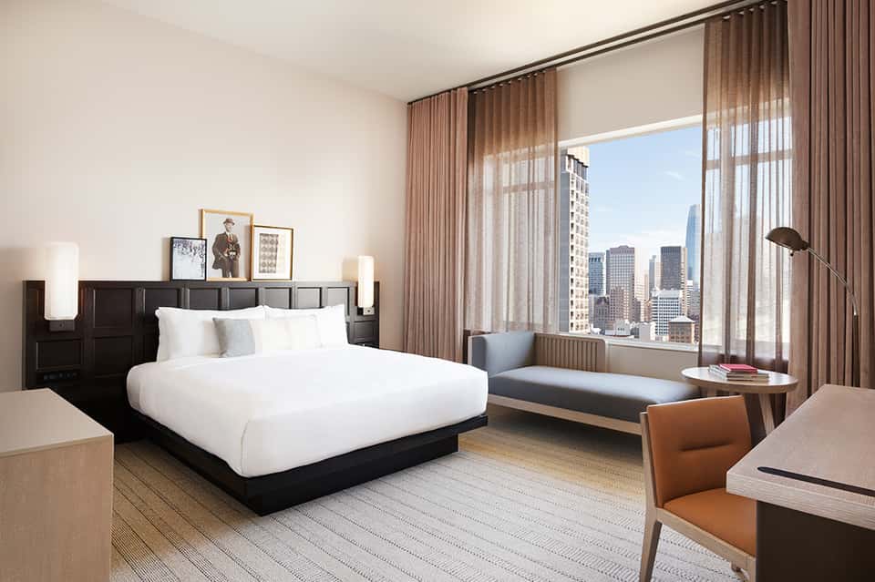Modern hotel bedroom with city skyline view, white bedding, dark headboard, and floor-to-ceiling windows