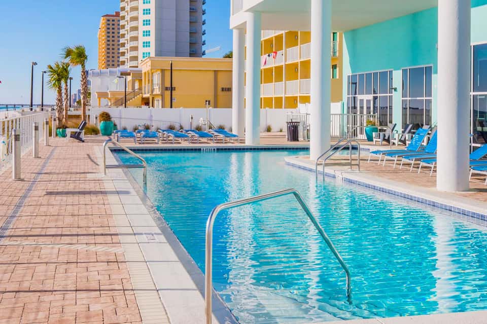 Bright turquoise pools with lounge chairs, colorful modern buildings, and palm trees at a beachfront resort