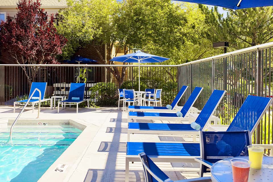 Resort pool area with blue lounge chairs, umbrellas, and manicured landscaping
