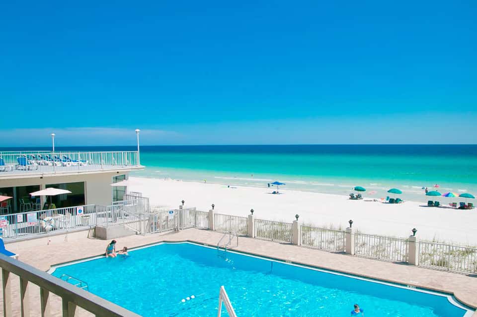 Beachfront hotel pool with turquoise water, white railing, and sandy beach with umbrellas beyond