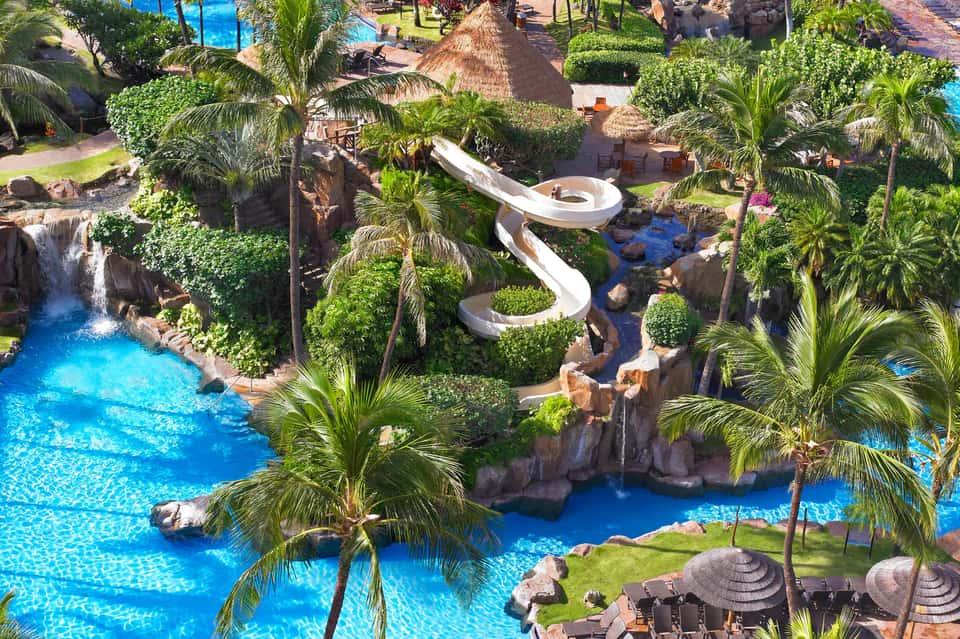 Aerial view of tropical resort with lazy river, waterslide, pools, lush landscaping, and palm trees