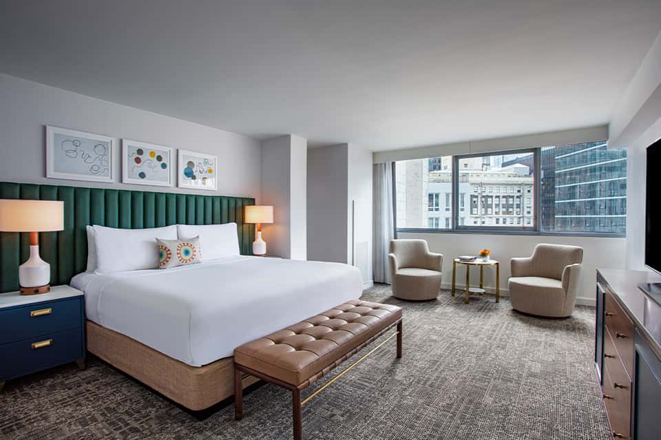 Modern hotel bedroom with green headboard, white bedding, city view windows, and contemporary furnishings